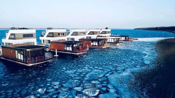 Conjunto de casas flotantes con terrazas abiertas y ventanas amplias, unidas por pasarelas, ubicadas sobre una masa de agua con formaciones de hielo visibles.