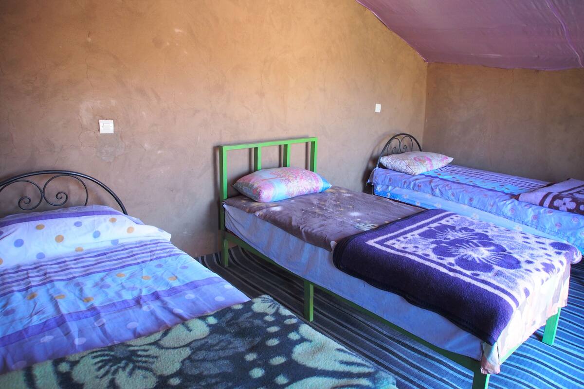 A room with three single beds, each with a patterned bedspread and pillows. The room has a sloped ceiling and a plain wall.