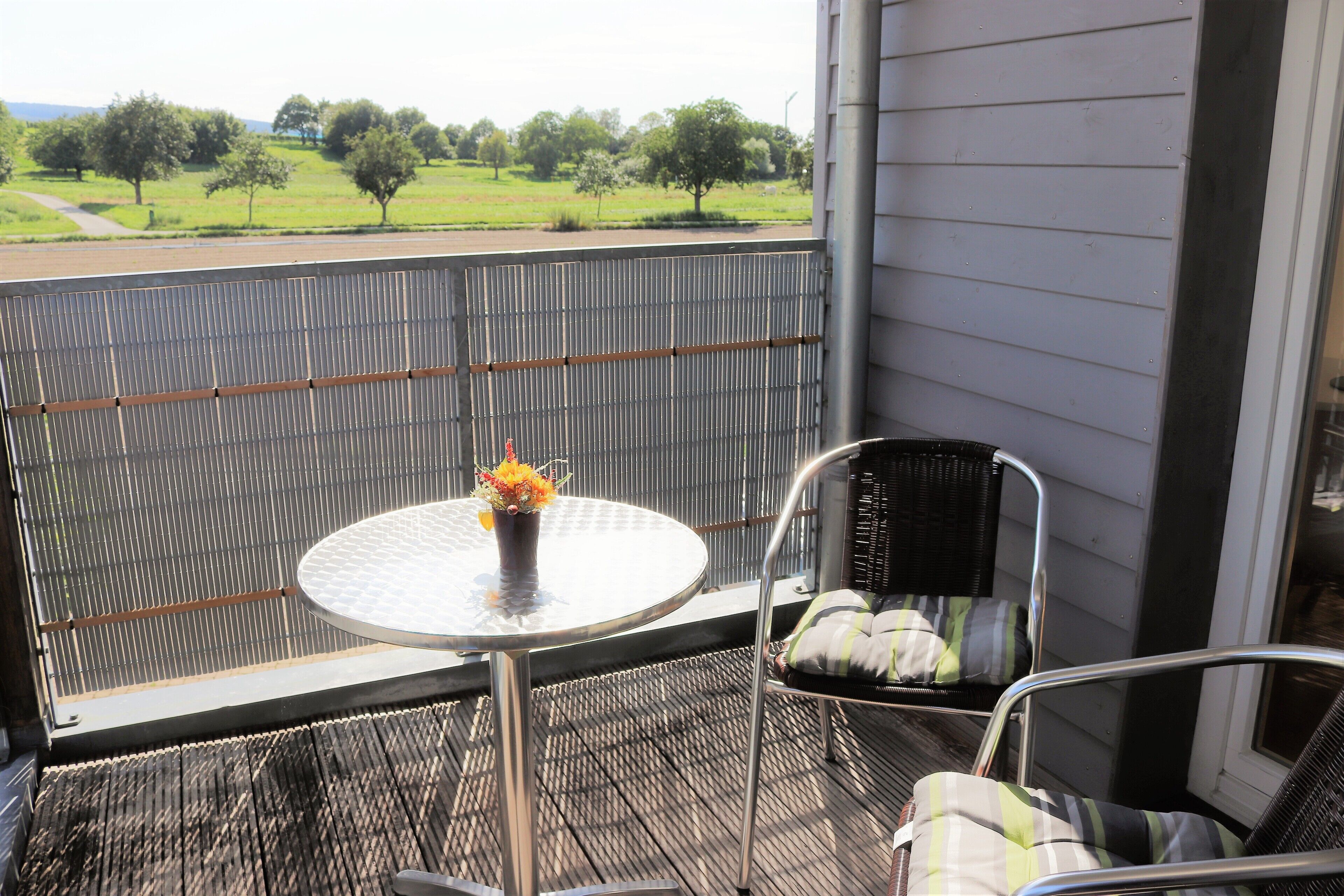 A balcony with a metal table and two wicker chairs, one with a cushion.