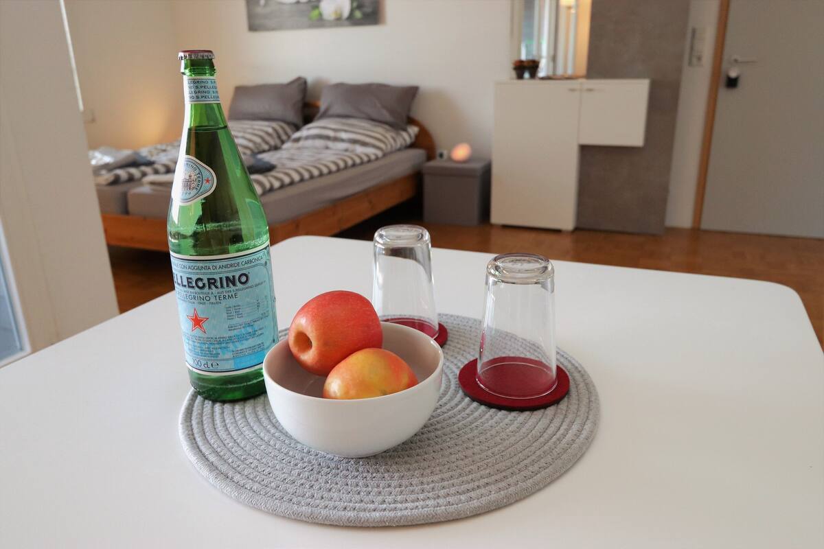 A bottle of Pellegrino water, a bowl with apples, and salt and pepper shakers on a table in a room with a bed and a nightstand.