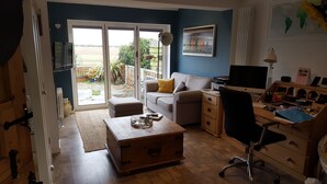 A cozy living room with a sofa, a wooden chest, a desk with a computer, and a view through a glass door to a garden. - B&B By The Sea (Lymington)