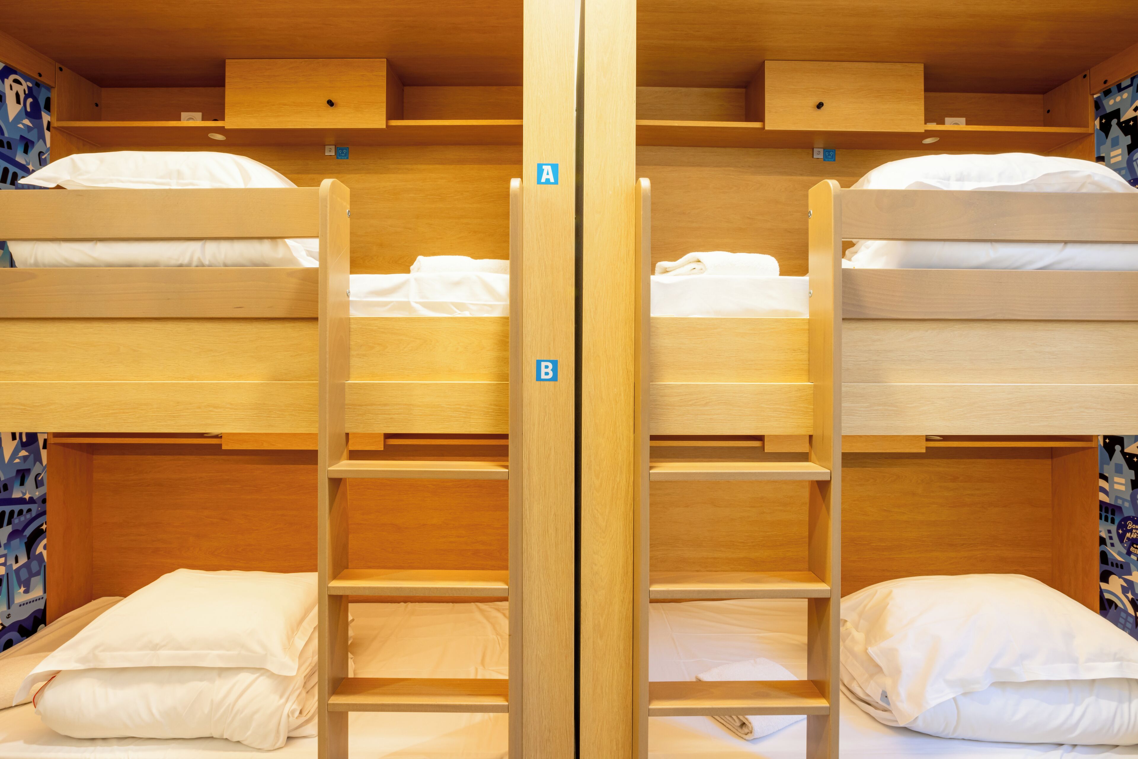 Une chambre avec des lits superposés en bois, chacun muni d’un oreiller et d’une petite étagère. Deux étiquettes, portant les lettres « A » et « B », sont apposées sur la cloison centrale.