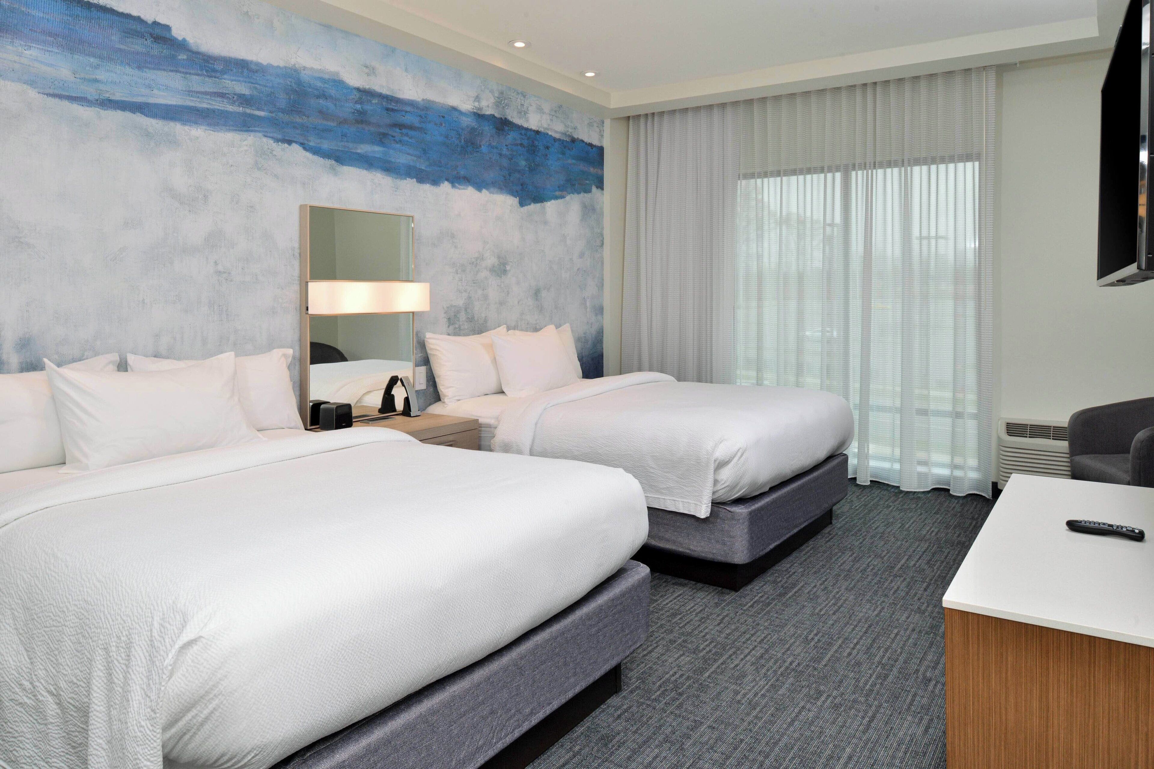 A hotel room with two beds, a desk, a chair, a TV, and a large window with curtains.