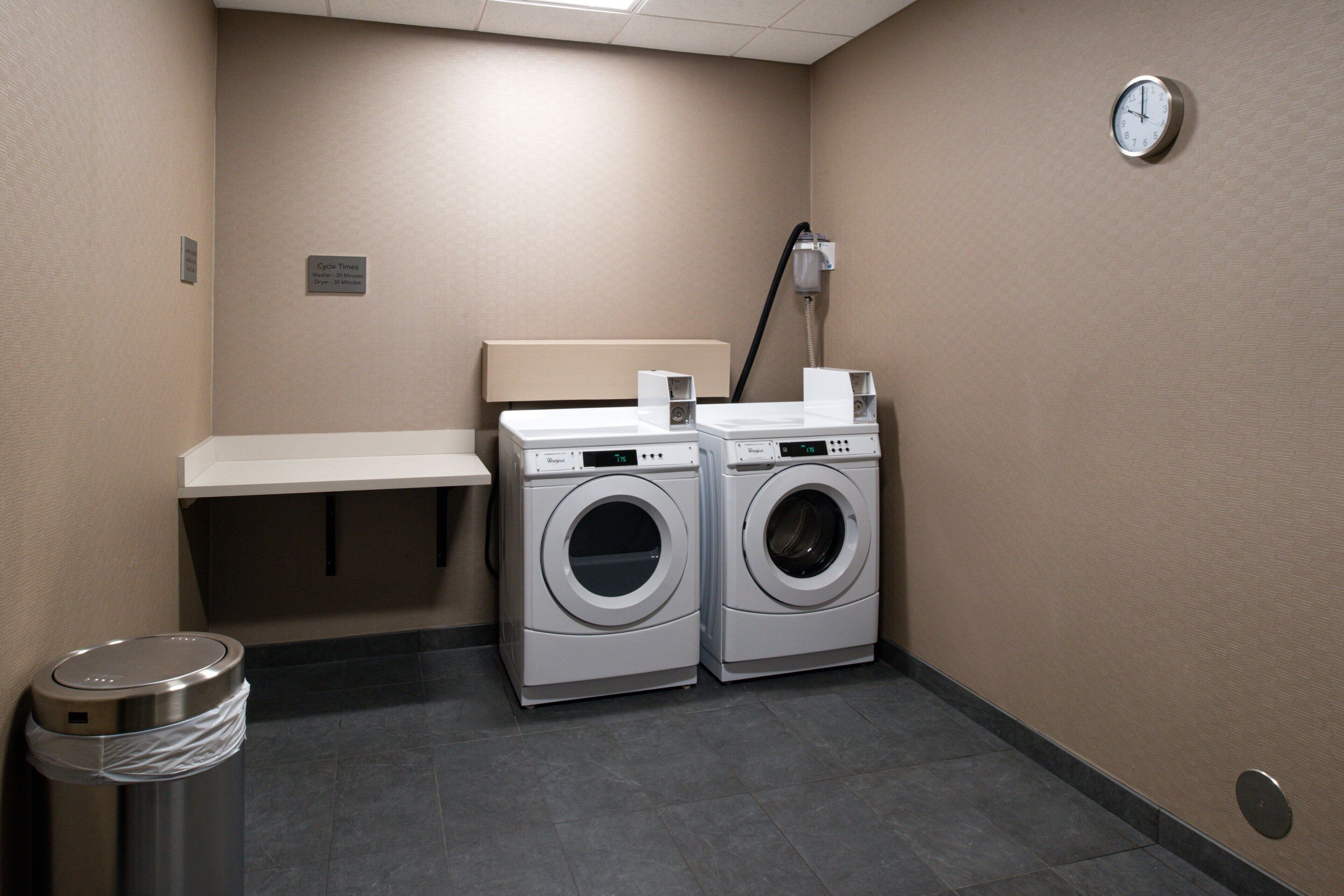A laundry room with two front-loading washing machines, a white bench, a trash can, and a wall-mounted clock.