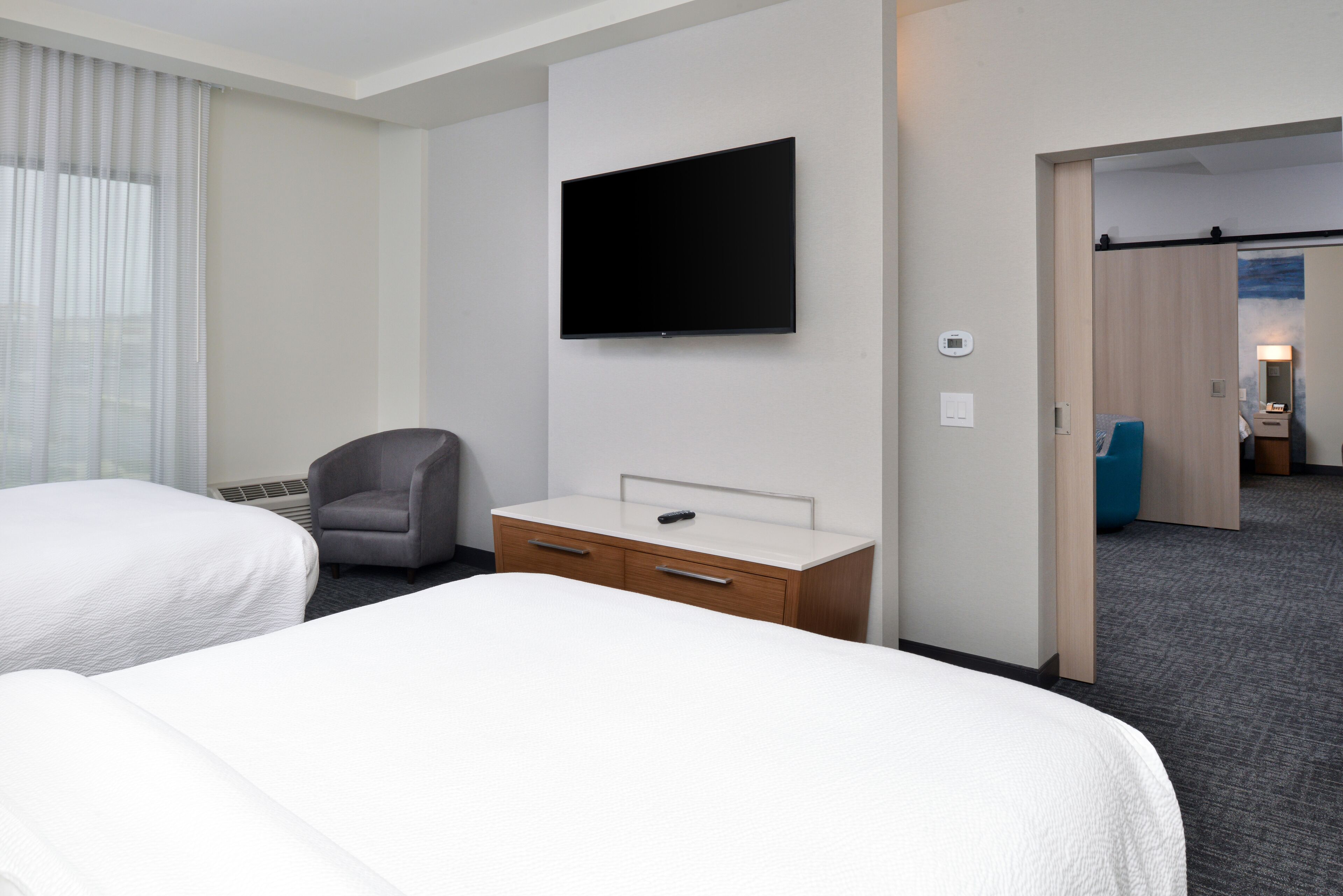 Et hotelværelse med to senge, et fladskærms-tv, et skrivebord med skuffer, en stol og en dør, der fører til et andet værelse.