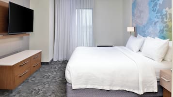 A hotel room with a bed, a flat-screen TV mounted on a wooden cabinet, a window with sheer curtains, and a colorful abstract wall mural.
