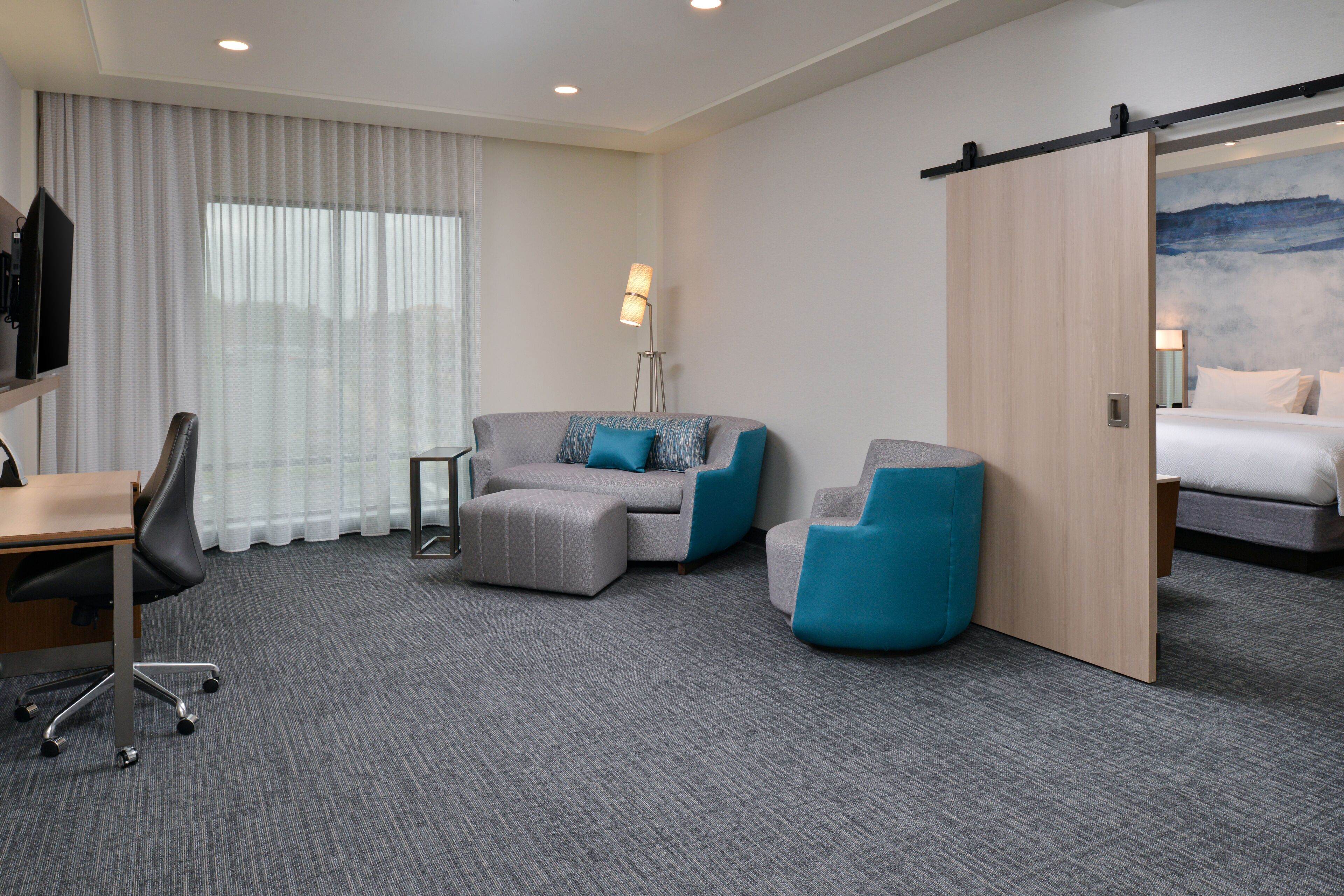 Une chambre d’hôtel équipée d’un bureau, de deux fauteuils, d’un canapé, d’un lit, d’une porte coulissante, d’une lampe et d’une grande fenêtre avec des rideaux.