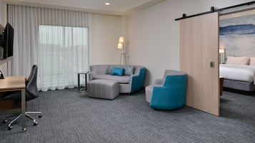 Une chambre d’hôtel équipée d’un bureau, de deux fauteuils, d’un canapé, d’un lit, d’une porte coulissante, d’une lampe et d’une grande fenêtre avec des rideaux.