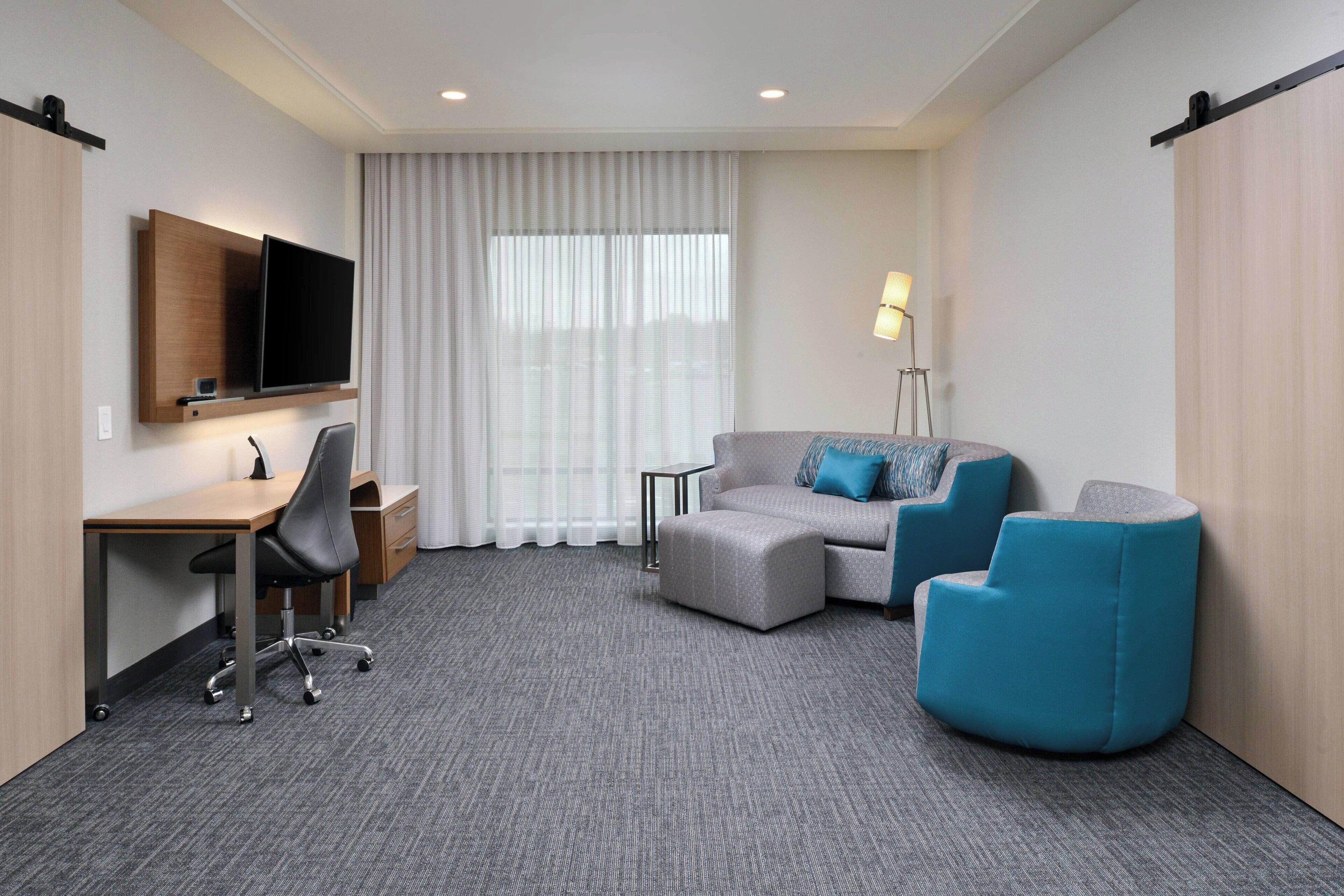 A hotel room with a desk, a flat-screen TV mounted on the wall, a sofa with blue cushions, and a standing lamp.