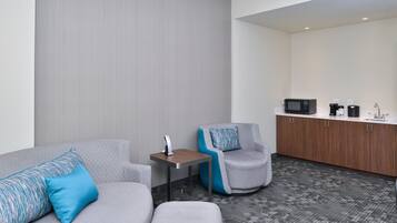 A modern hotel room with a gray sofa, a gray armchair, a small wooden table, and a kitchenette with a microwave, coffee maker, and cabinets.