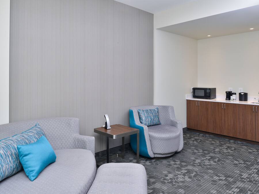 A modern hotel room with a gray sofa, a gray armchair, a small wooden table, and a kitchenette with a microwave, coffee maker, and cabinets.