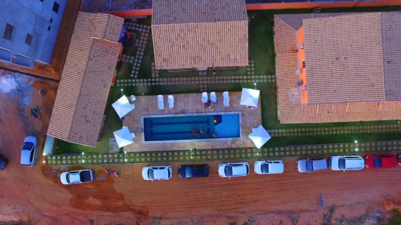 An aerial view of a residential area with a swimming pool surrounded by lounge chairs and umbrellas, a parking lot with cars, and houses with similar roof designs.