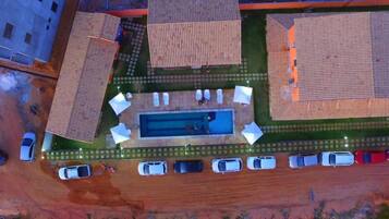 An aerial view of a residential area with a swimming pool surrounded by lounge chairs and umbrellas, a parking lot with cars, and houses with similar roof designs.