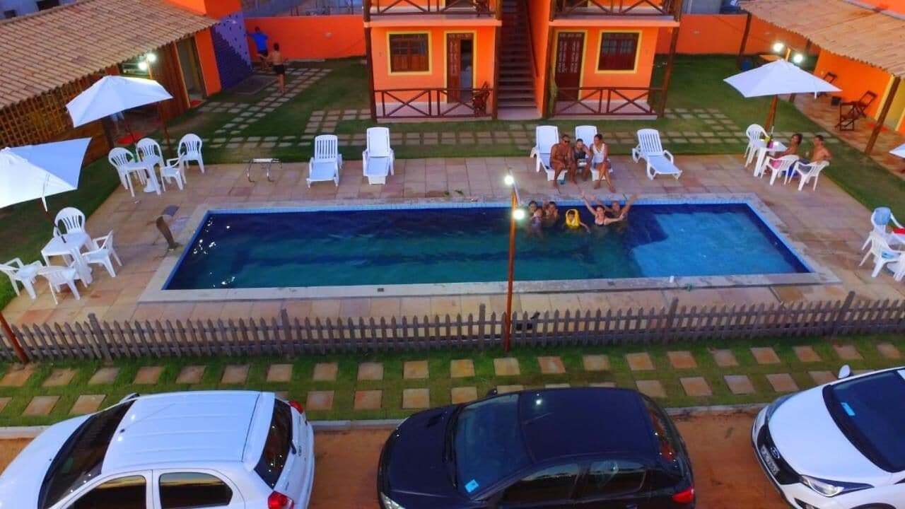 En swimmingpool omgivet af hvide stole og parasoller, med et træhegn og en bygning i baggrunden.