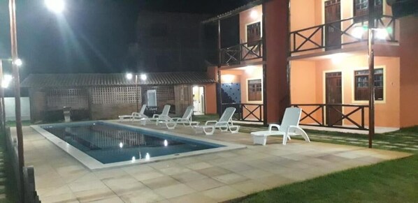Una piscina con sillones de descanso, un porche cubierto y un edificio de dos plantas con balcones.