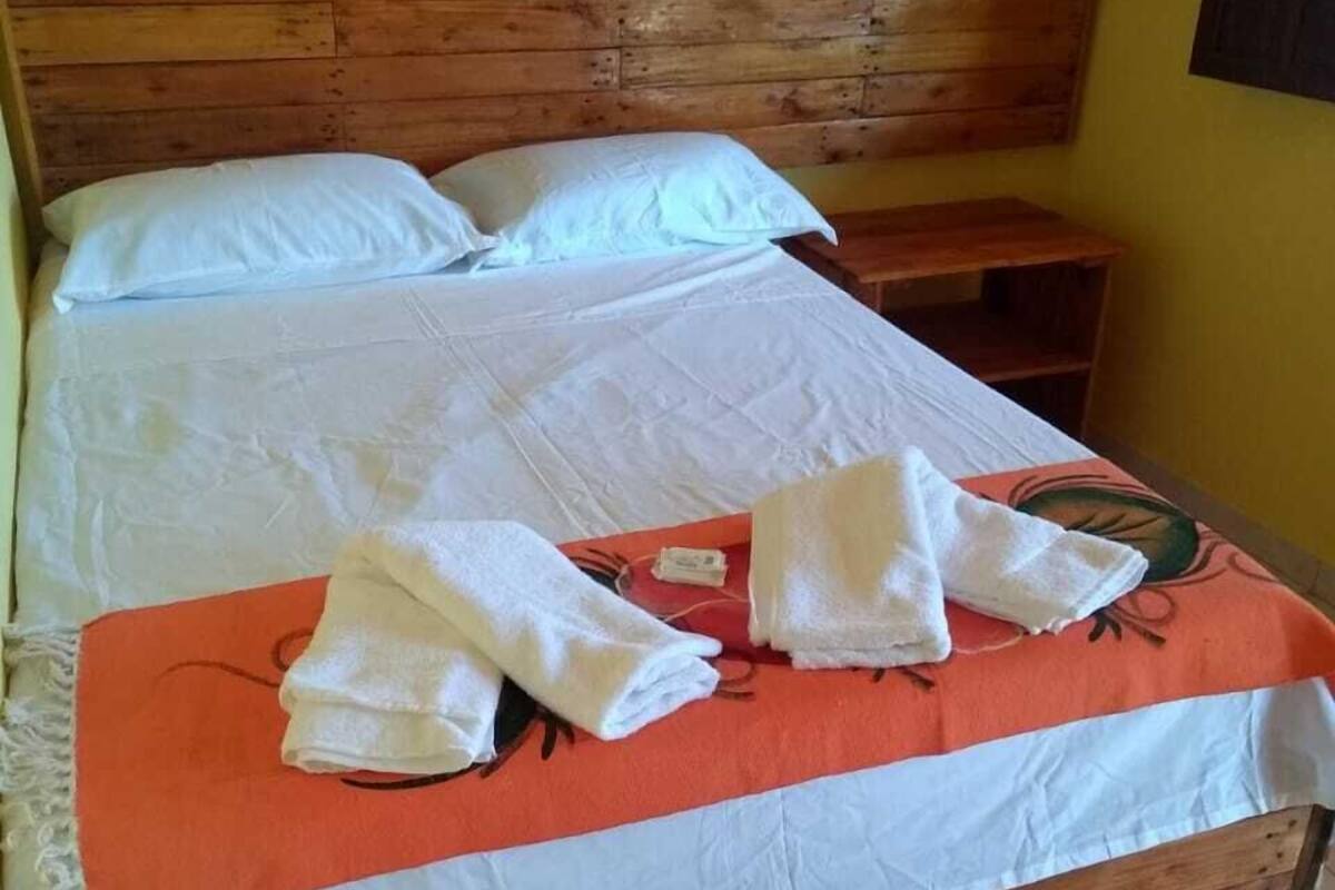 Una cama de madera con sábanas blancas y una manta naranja, una mesita de noche de madera, y una alfombra roja y blanca con estampado sobre un piso de baldosas.