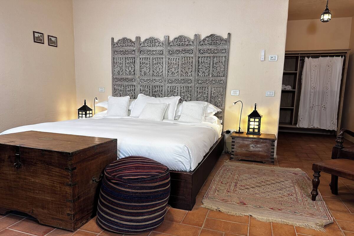 Un dormitorio con una cama grande, un cofre de madera, un reposapiés a rayas, una alfombra estampada y un cabecero decorativo.