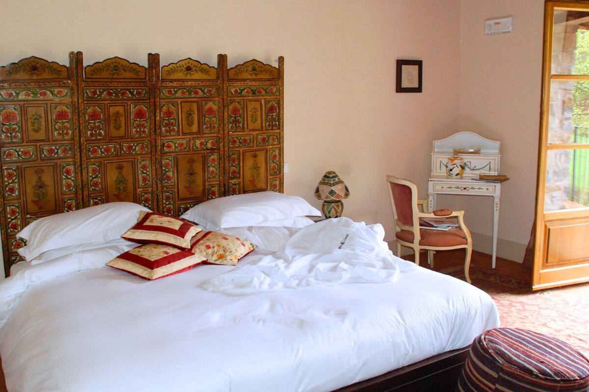 Una cama con sábanas blancas, cabecera de madera con motivos florales y un pequeño escritorio con silla.