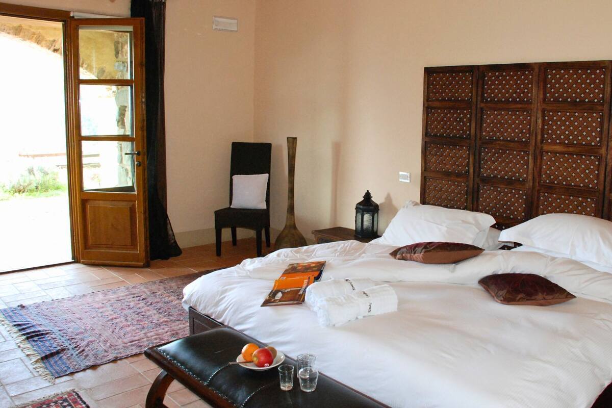 Un dormitorio con una cama grande, un cabecero de madera, una silla, una mesita de noche y una pequeña mesa con fruta y vasos.
