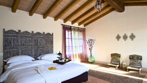 A bedroom with a large bed, wooden headboard, a chandelier, a window with curtains, a vase with dried flowers, and two chairs. - Podere Conti (Filattiera)