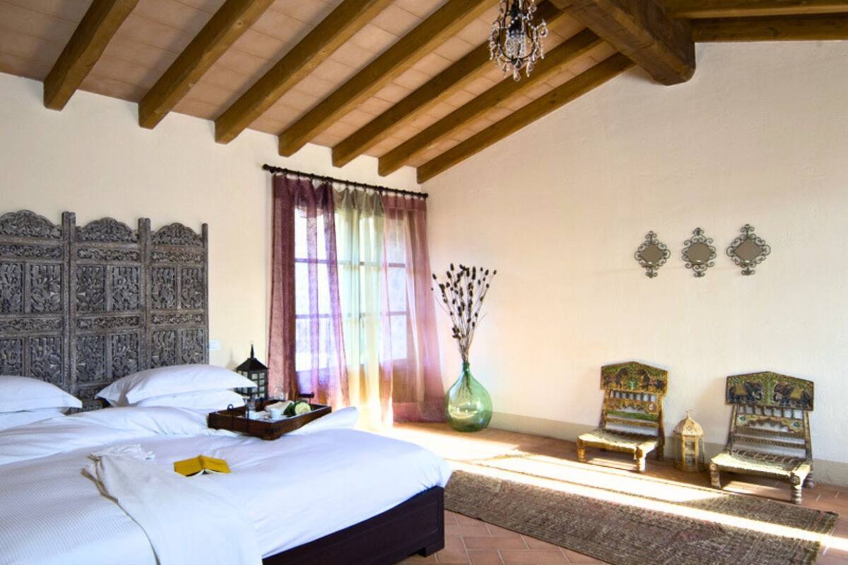 Un dormitorio con una cama grande, cabecera de madera, candelabro, ventana con cortinas, un jarrón con flores secas y dos sillones.
