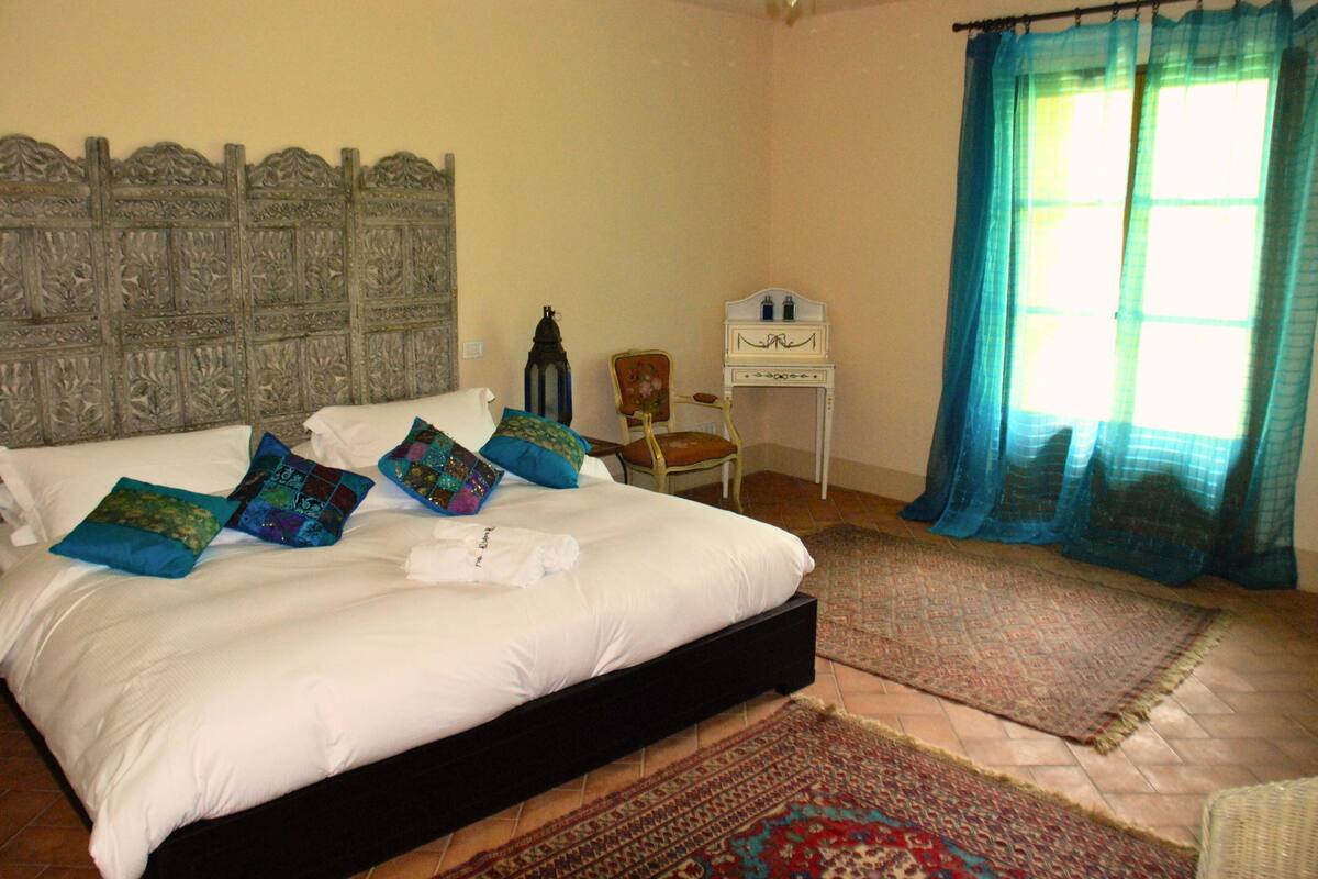 Un dormitorio con una cama grande, un cabecero, una silla, una mesita y una ventana con cortinas.