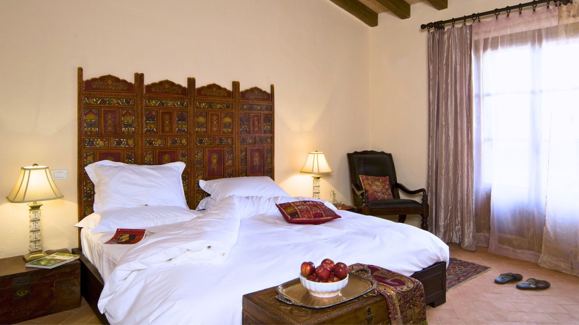 A neatly made bed with white linens, a decorative headboard, a bedside table with a lamp, a chair, and a window with curtains.