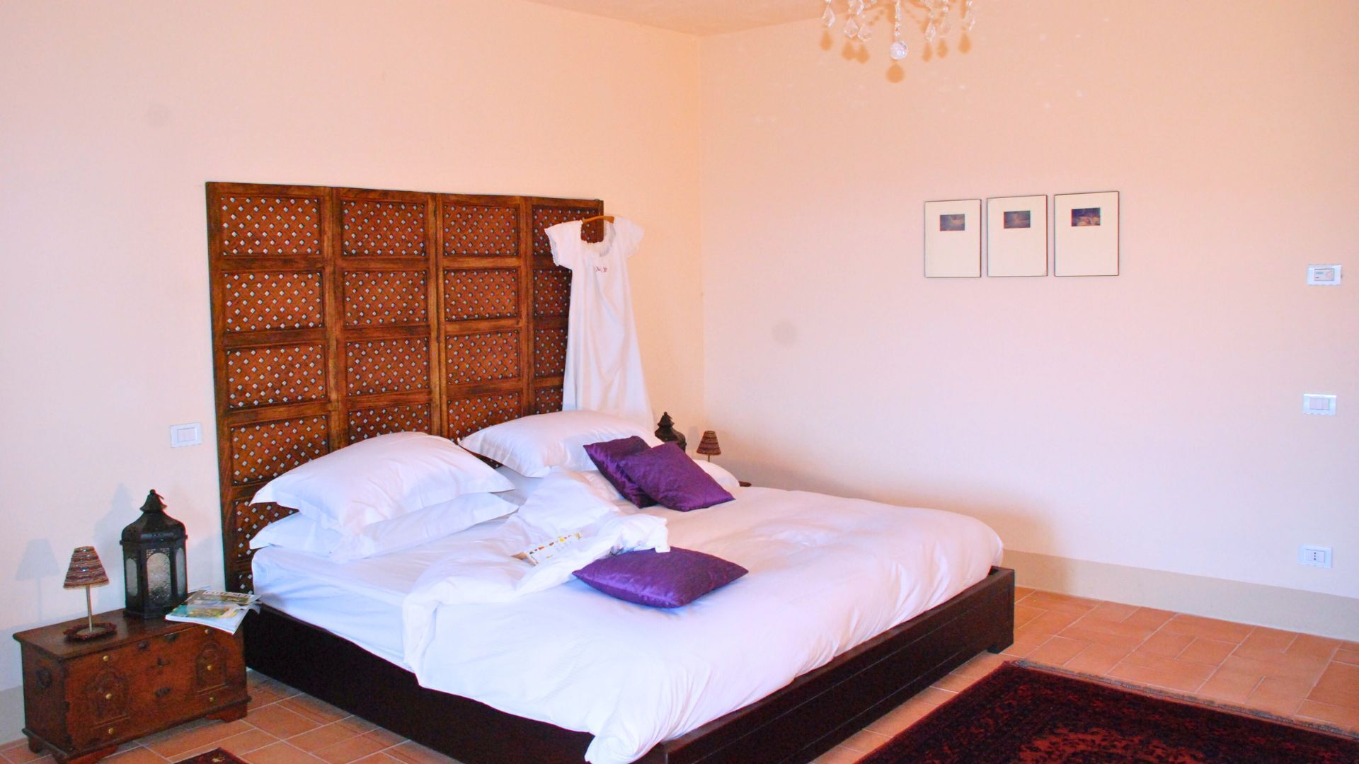 A bedroom with a wooden headboard, a bed with white and purple bedding, a nightstand with a lamp, and a red rug.