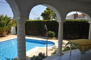 Uma piscina com estrutura de coluna branca, uma mesa com toalha amarela e um sebe ao fundo.