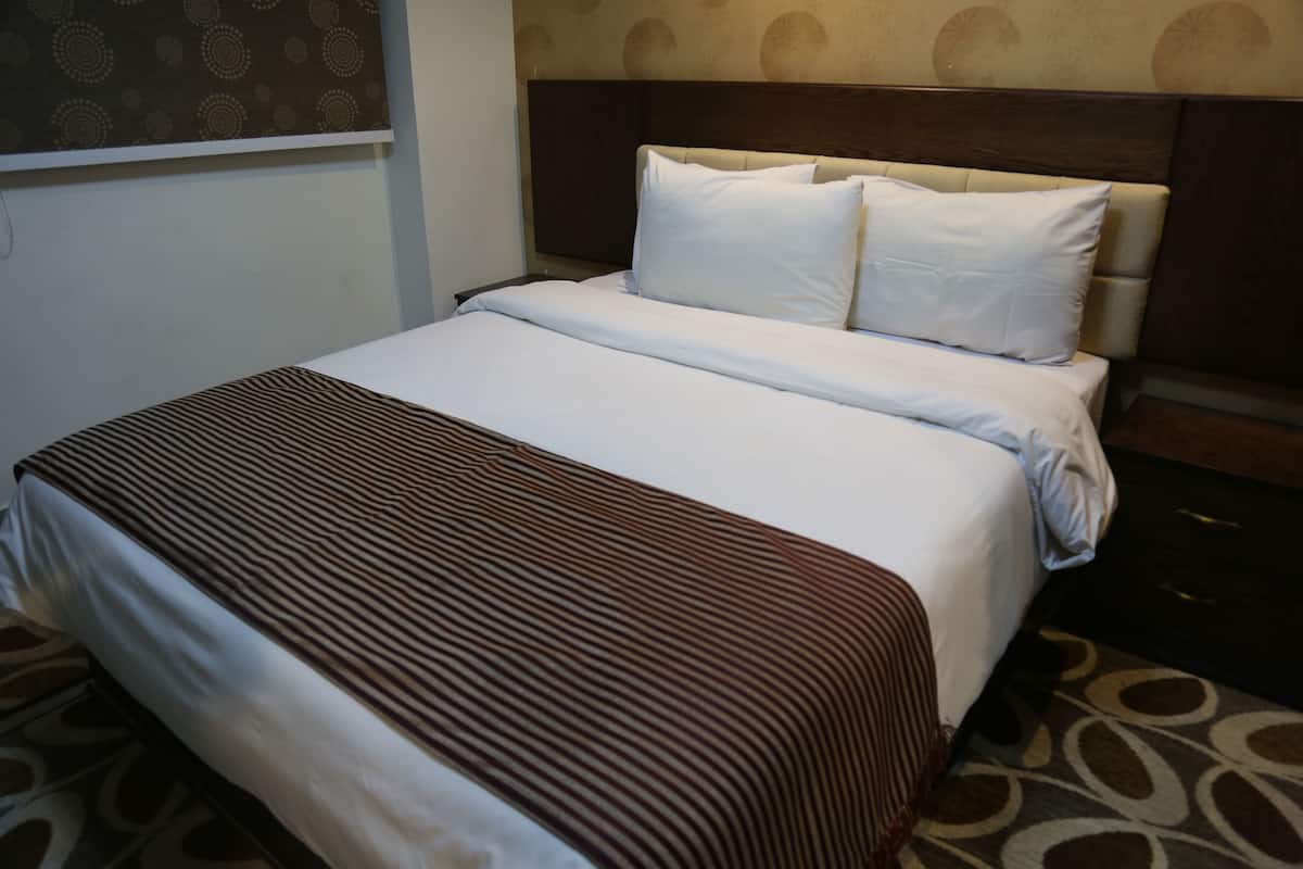 A hotel room with a bed, a headboard, a window with a patterned curtain, and a patterned carpet.