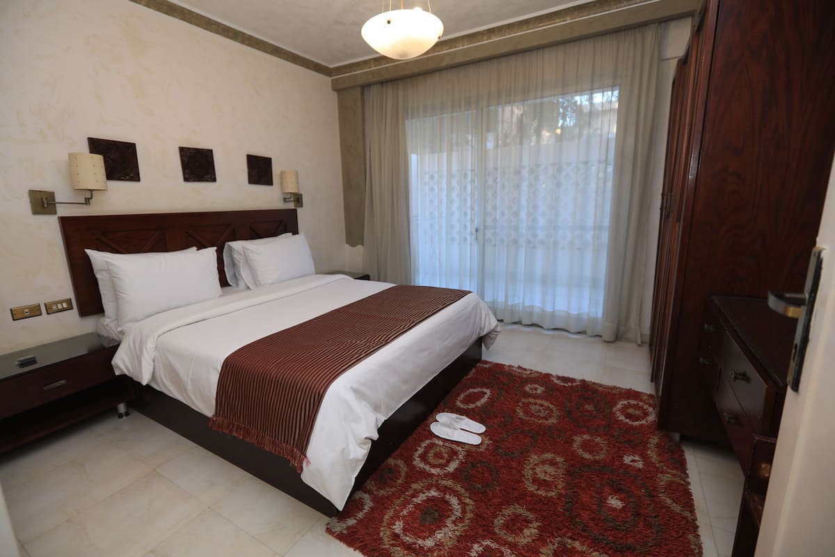 A hotel room with a large bed, a wooden dresser, a red patterned rug, and a window with sheer curtains.