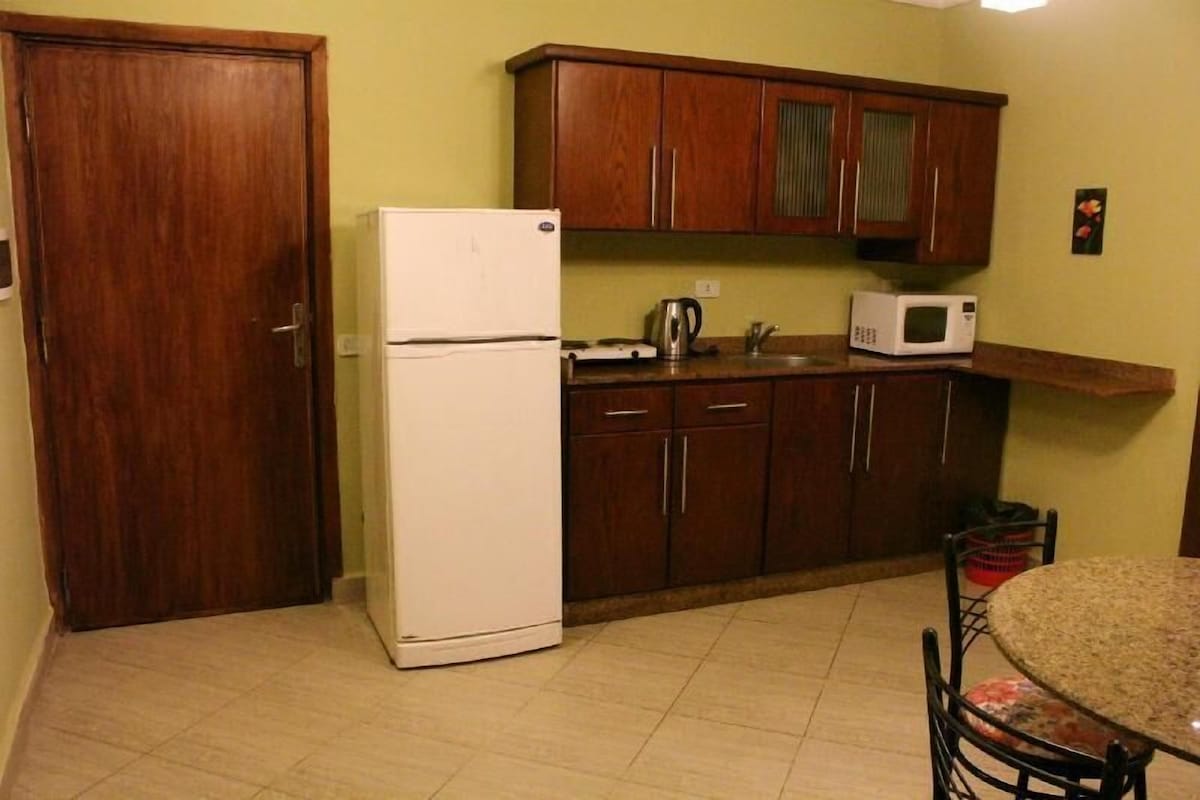 A compact kitchen with wooden cabinets, a white refrigerator, a microwave, and a small dining table with chairs.