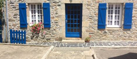 A stone building with a tiled entrance, blue shutters, and a small window with a grid pattern.