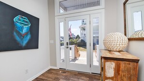 A room with a wooden floor, a painting of a boat, a wooden cabinet, and a view through a glass door to a balcony with a railing and an American flag. - Oceanside, Private Pool, Close to Beach, Hot Tub! SP2, Blew Bayou (Duck)