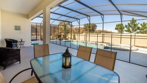 Un patio techado con una mesa de vidrio, sillas de mimbre y un farol. Al fondo, se observa una piscina y un jacuzzi.