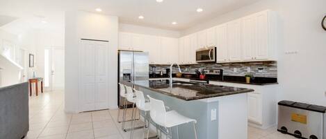 A modern kitchen with white cabinets, a central island with bar stools, stainless steel appliances, and a tiled floor.