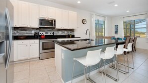 Cocina moderna con armarios blancos, electrodomésticos de acero inoxidable, encimera de granito y una zona de bar con taburetes blancos.