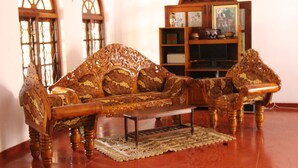 A traditional wooden sofa set with intricate carvings, a matching armchair, and a small wooden coffee table in a room with wooden shelving and a decorative mirror.