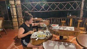 A woman dining at a rustic outdoor restaurant with wooden tables and chairs, surrounded by bamboo structures and a decorative metal fence.