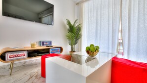 Ein modernes Wohnzimmer mit einem Glastisch, einem roten Sofa, einem an der Wand befestigten Fernseher und einem Monopoly-Spiel auf einem Holztischchen.