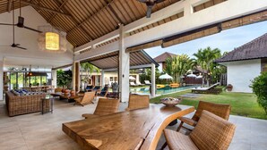 Un amplio espacio al aire libre con una gran mesa de madera, sillas de mimbre, una piscina y una zona de estar cubierta con sombrillas.