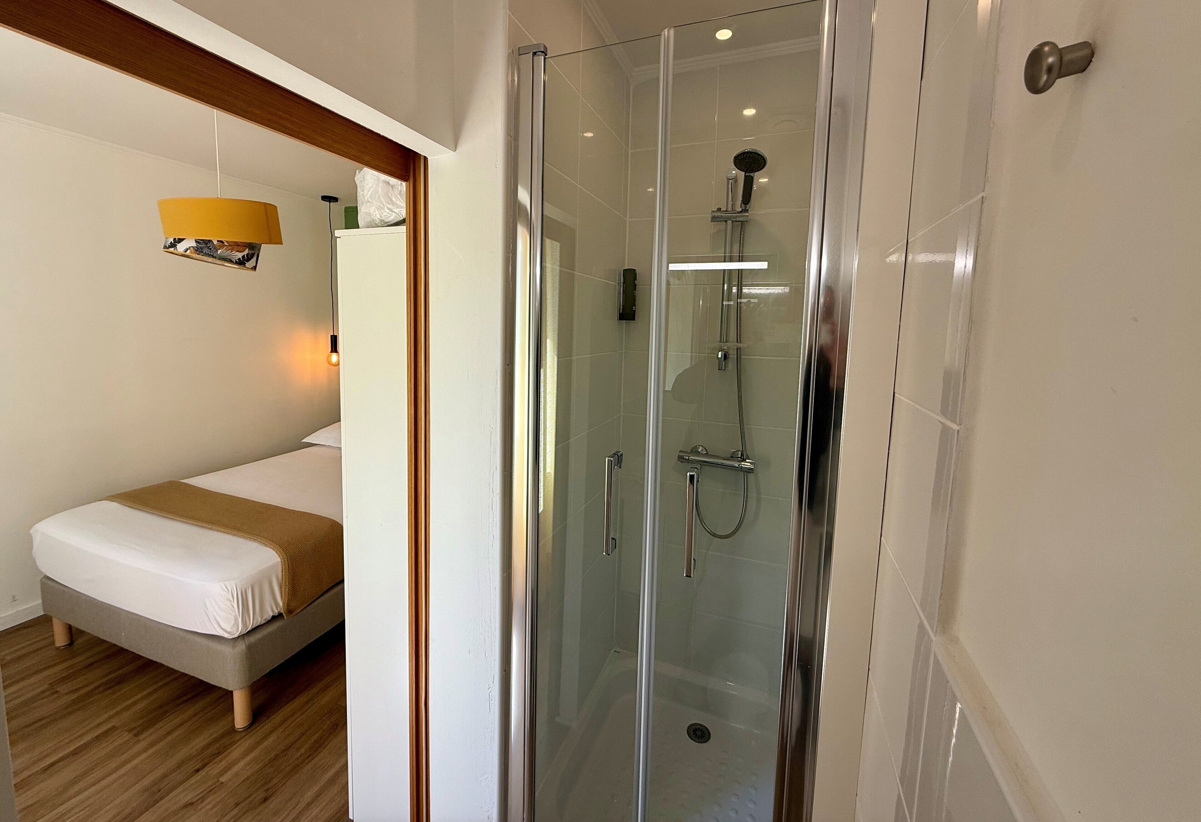 Une chambre d’hôtel dotée d’une douche en verre, d’un lit recouvert d’une courtepointe blanche et marron, et d’un plancher en bois.