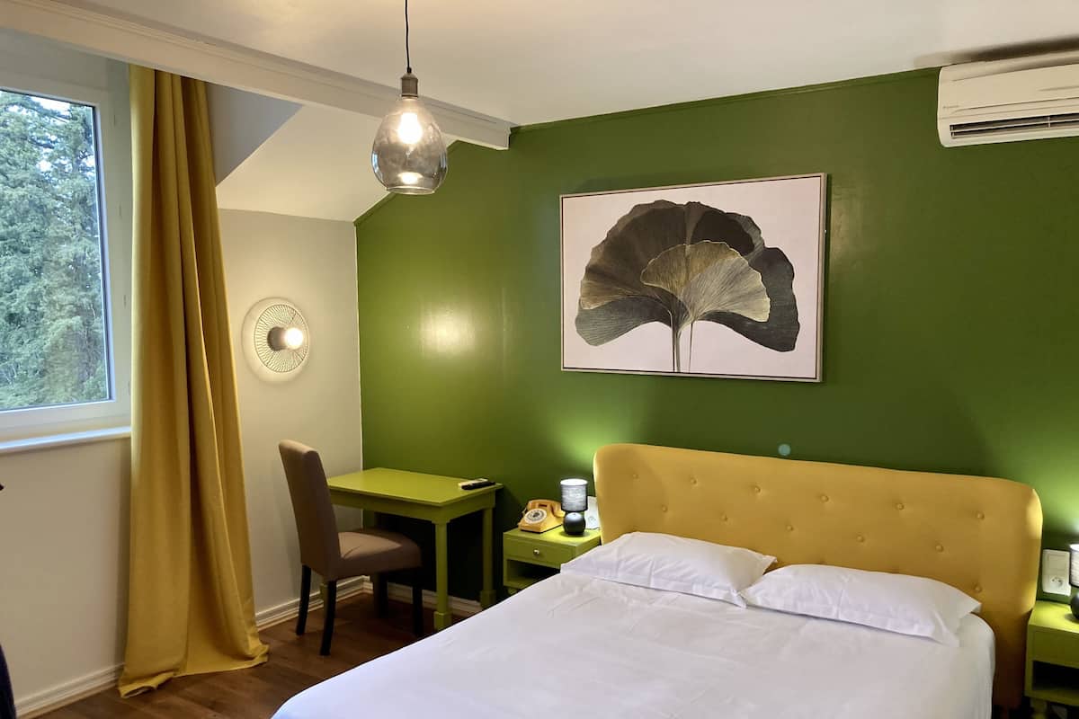 A hotel room with a bed, a desk, a chair, a lamp, and a large leaf artwork on the wall.
