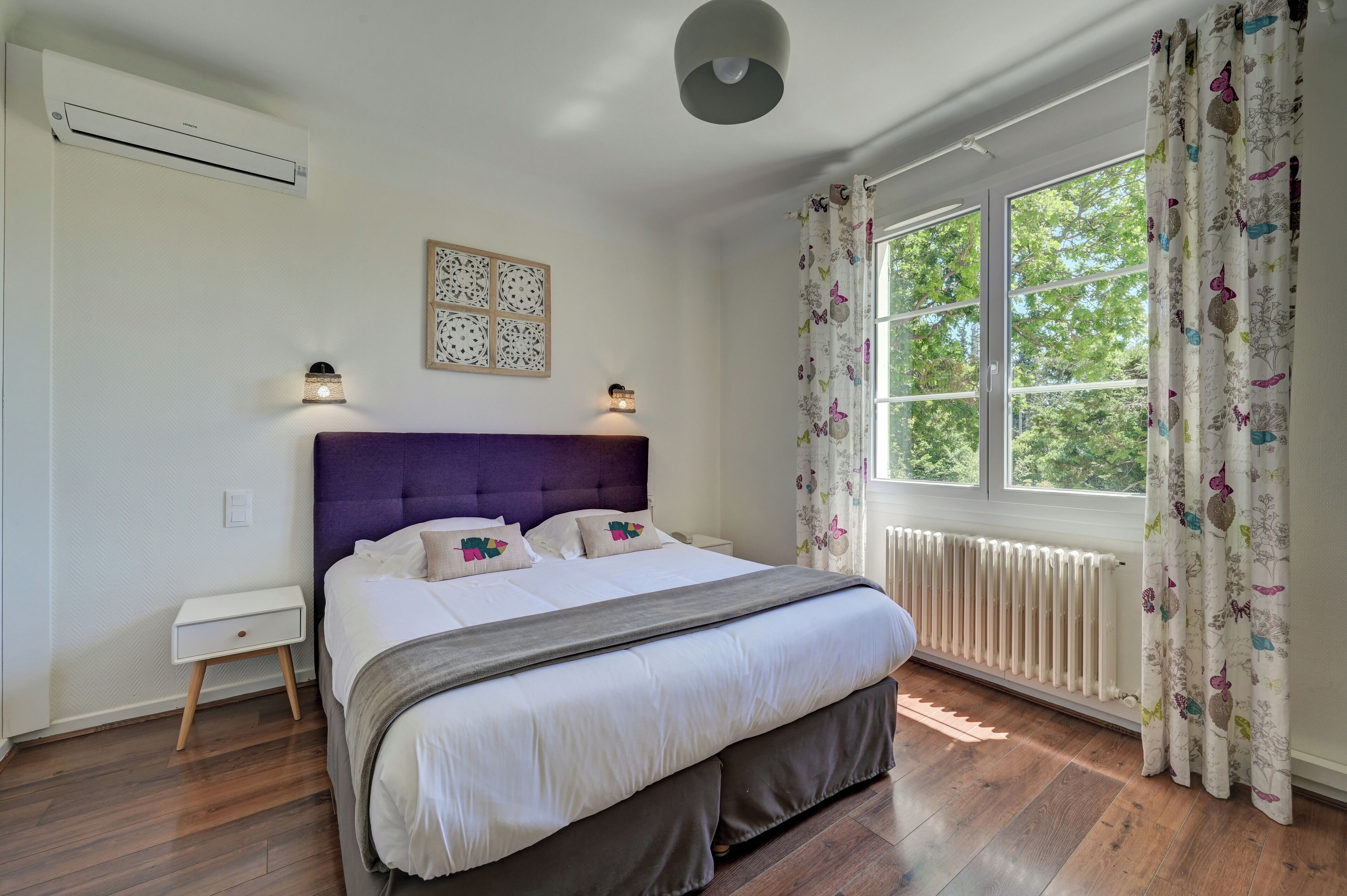 Un dormitorio con una cama grande, una mesita de noche, aire acondicionado en la pared, una lámpara en el techo, una ventana con cortinas y un radiador.