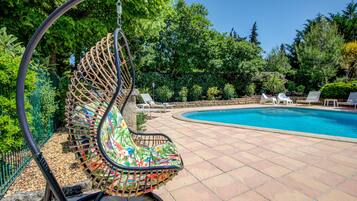 A hanging chair with a colorful cushion by a swimming pool surrounded by greenery.
