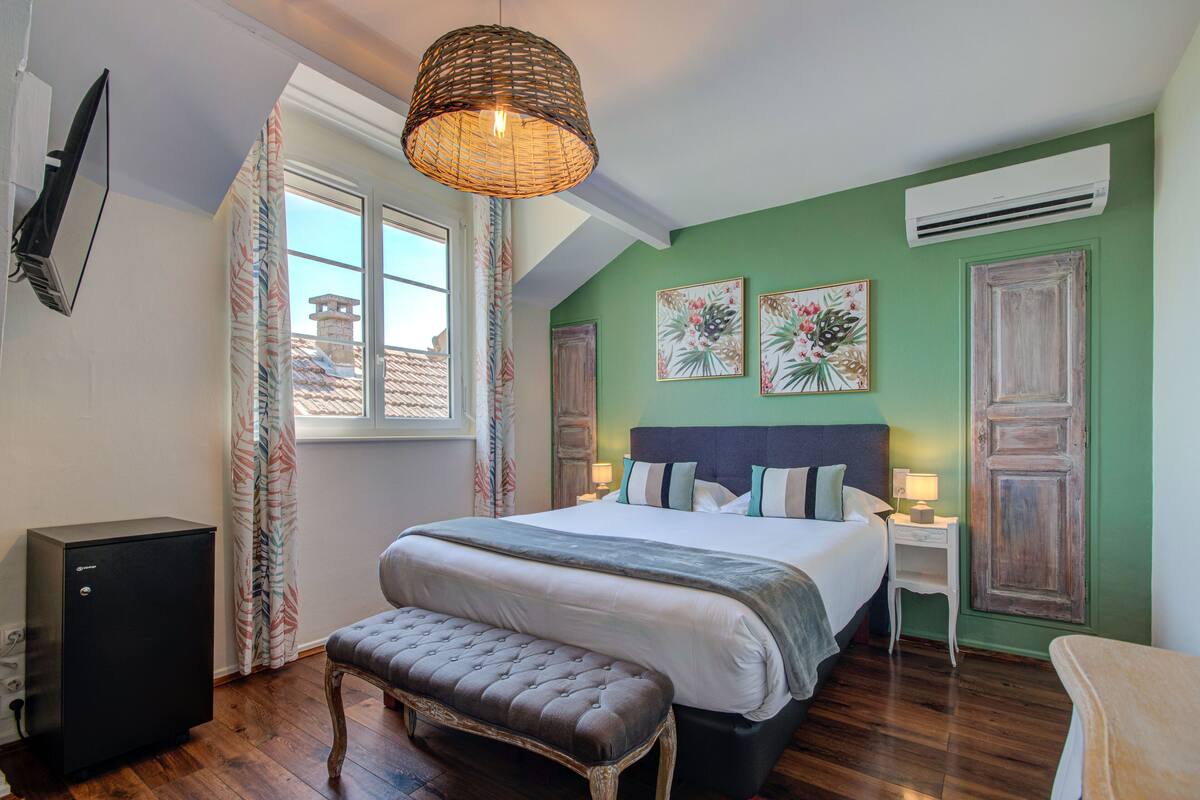A bedroom with a bed, a bench, a small table, an air conditioner, and a window with curtains.
