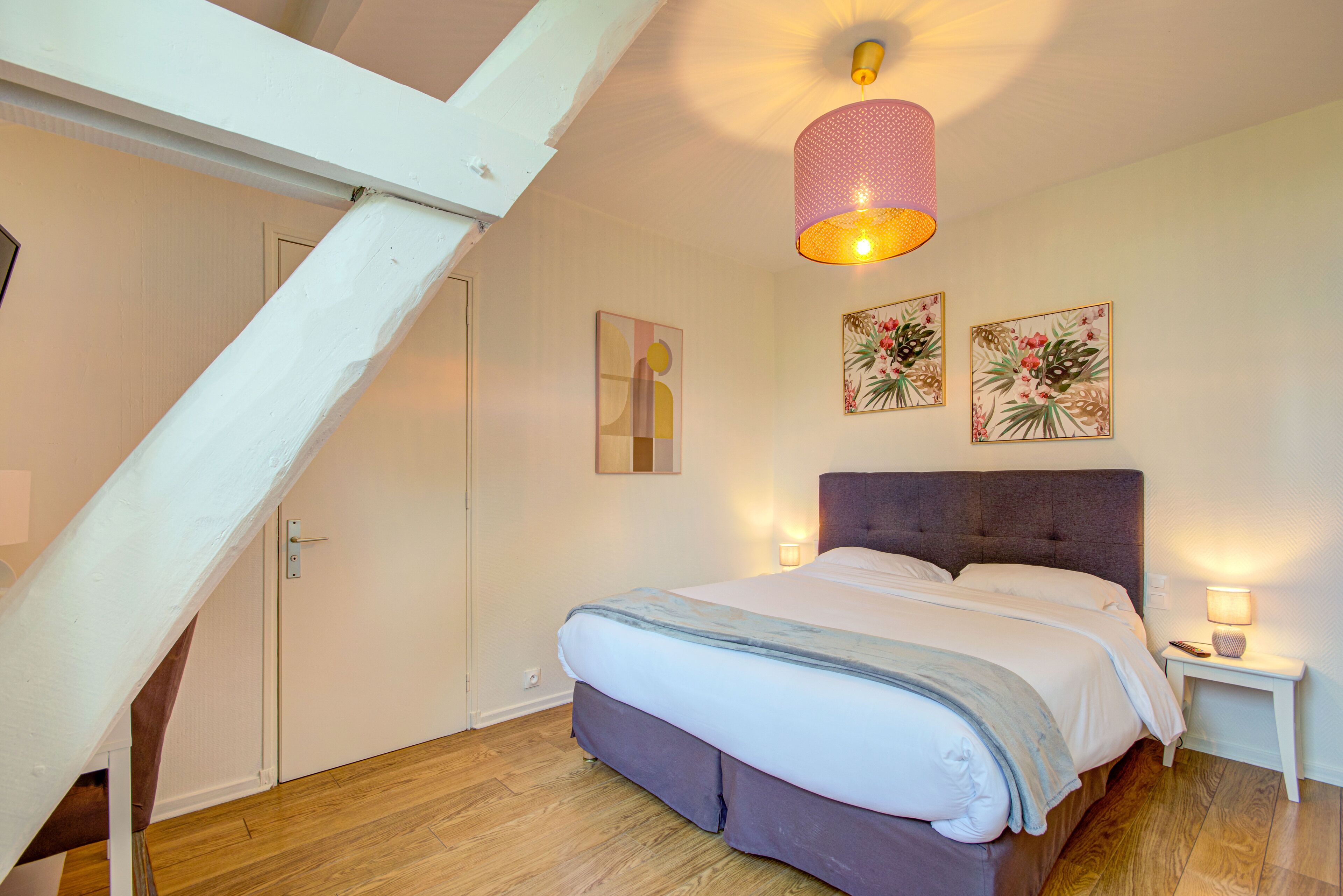 A bedroom with a bed, a nightstand, a wall-mounted lamp, and two framed pictures.