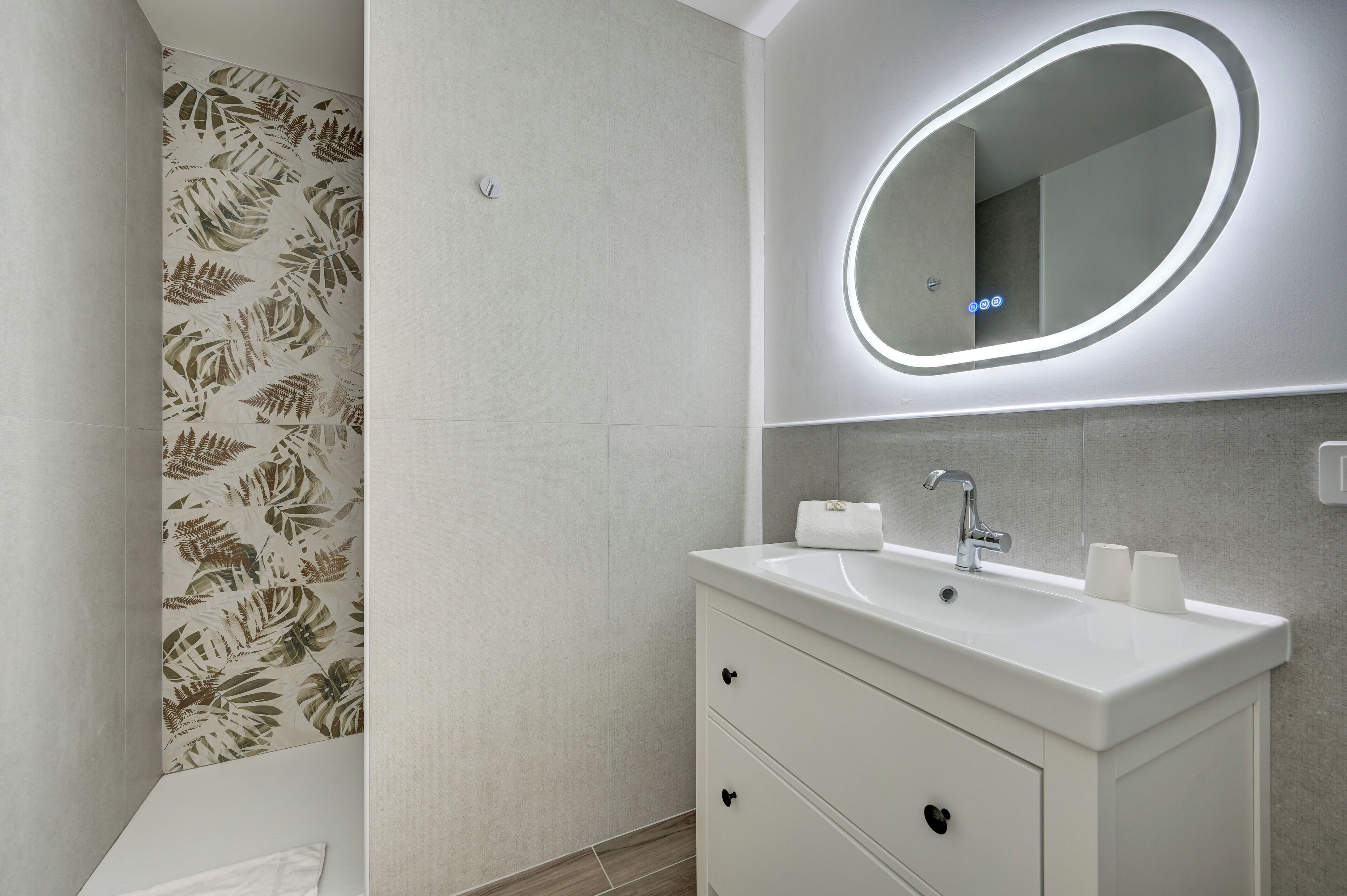 Een moderne badkamer met een witte wastafel, een ronde spiegel en een douche met behang met een bladmotief.