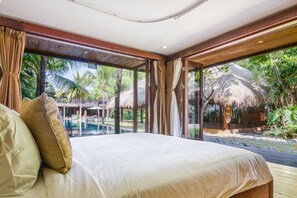 A bedroom with a large bed, a view of a pool, and a thatched-roof structure outside.