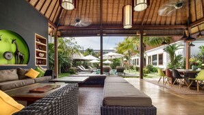 Amplia sala de estar con un sofá grande, una mesa de centro de mimbre y una mesa de comedor con sillas. Se ve un área de piscina a través de puertas de vidrio.